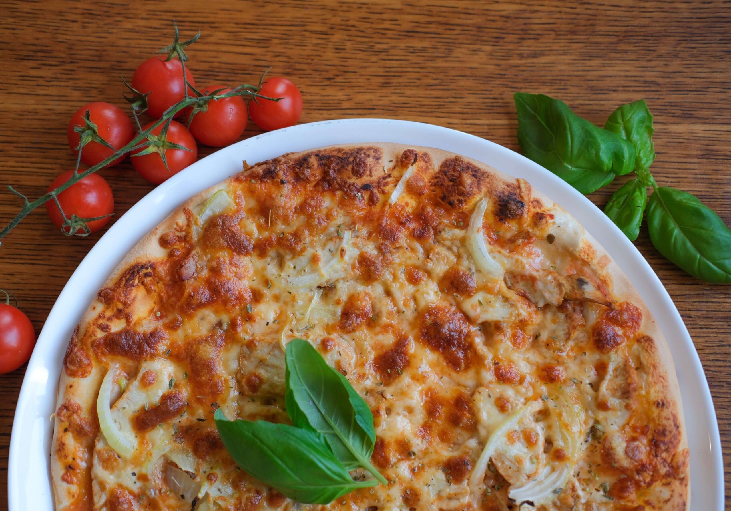 Pizza z serem, kurczakiem i cebulą, ozdobiona listkami bazylii, podana na białym talerzu, obok świeże pomidorki koktajlowe.