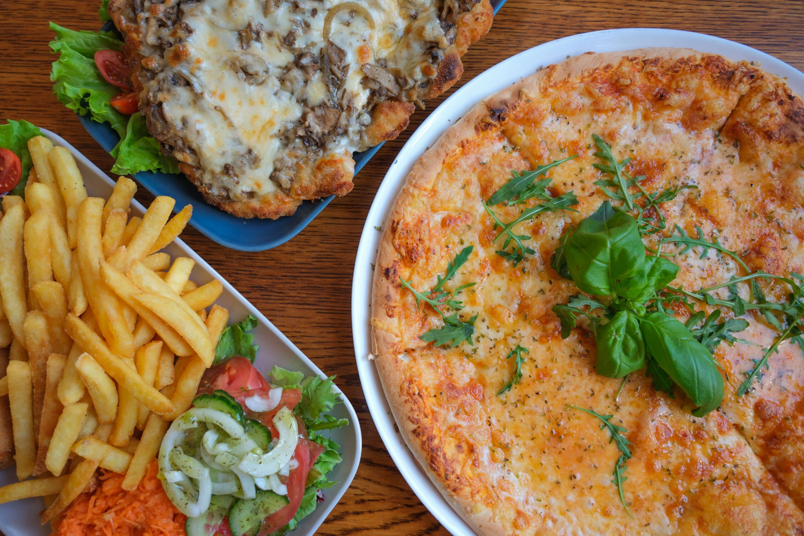 Pizza z serem i ziołami, panierowany kotlet z pieczarkami i serem oraz porcja frytek z surówką na drewnianym stole